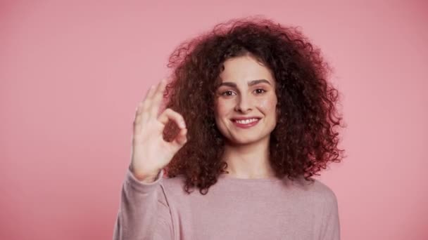 Vencedor. Sucesso. Menina positiva fazendo OK assinar sobre fundo rosa e sorri para a câmera. Linguagem corporal. Jovem mulher encaracolada com moderno brilho sardas maquiagem — Vídeo de Stock