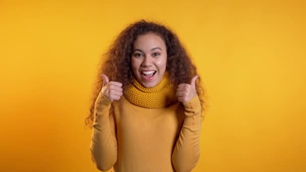 Winnaar. Succes. Positief meisje maakt duimen omhoog teken over gele achtergrond en glimlacht naar camera. Lichaamstaal. jong gemengd ras krullend vrouw — Stockvideo