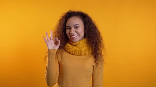 Chica positiva haciendo signo OK sobre fondo amarillo y sonríe a la cámara. El lenguaje corporal. Joven mujer rizada en total look cálido — Vídeo de stock