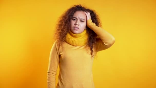 Young upset woman with curly hair having headache, studio portrait. Girl putting hands on head, isolated on yellow background. Concept of problems and headache. — Stock Video