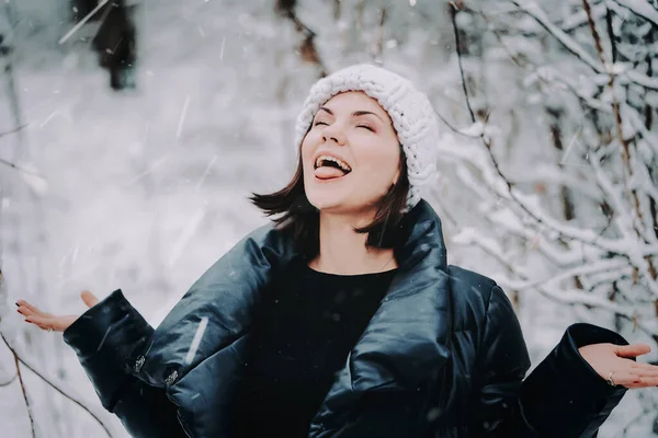 Nice girl in winter clothes in the forest catches snowflakes with her mouth — ストック写真