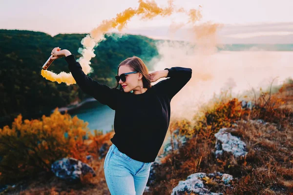 Giovane bella donna con bomba fumogena arancione o granata in drammatica illuminazione lunatica. Ragazza in felpa nera su una scogliera sopra il fiume — Foto Stock