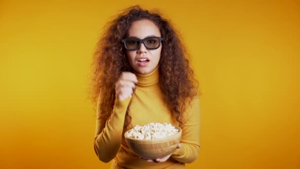 Jeune femme dans des lunettes 3D regarder un film et manger du pop-corn sur fond de studio jaune . — Video