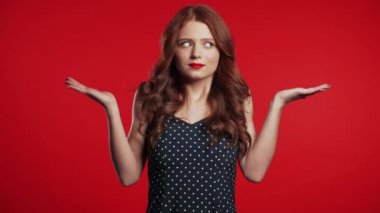  Young unsure girl shrugs her arms, makes gesture of I dont know, care, cant help anything .Young millenial woman on red background