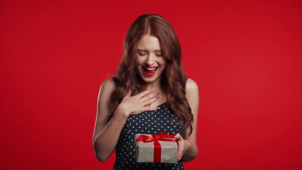 Joyful beautiful woman with perfect makeup holding gift box with bow on red wall background. Retro styled girl smiling, she is glad to get present. — Stockvideo