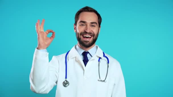 Portret van een lachende positieve dokter in professionele medische witte jas met OK teken gebaar. Doc geïsoleerd op blauwe achtergrond. — Stockvideo