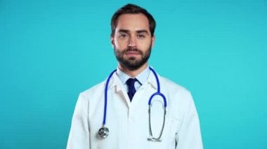 Profesyonel medikal beyaz önlüklü genç bir adamın portresi mavi stüdyo arka planında izole edilmiştir. Sakallı ve steteskoplu doktor.