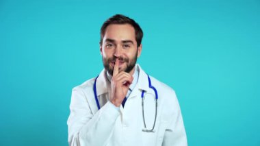 Profesyonel medikal beyaz önlüklü gülümseyen doktorun portresi mavi arka planda parmaklarını tutuyor. Şşş, sır, sessizlik jesti. Vücut dili. Doktor bey..