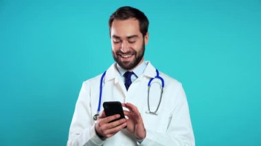 Mavi stüdyo arka planında izole edilmiş cep telefonu kullanan profesyonel beyaz önlüklü yakışıklı bir adamın portresi. Sakallı ve steteskoplu bir doktor. Modern teknoloji, internet kavramı.