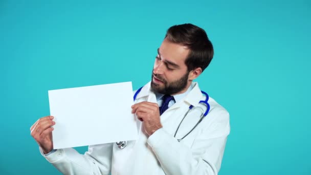 Portrait of young handsome man in professional medical white coat holding white horizontal a4 paper isolated on blue studio background. Doctor with beard and stethoscope. Copy space. — Stock Video