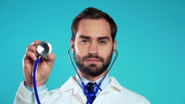 Portrait de jeune homme séduisant en manteau médical blanc professionnel utilisant un stéthoscope isolé sur fond bleu studio. L'homme pointe son instrument vers la caméra comme s'il écoutait un patient malade — Video