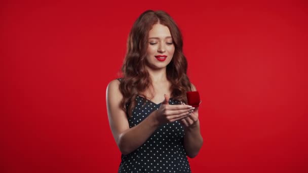 Mujer joven bastante europea en vestido de lunares sosteniendo pequeña caja de joyería con propuesta de matrimonio sobre fondo rojo. Chica disgustada decepcionada, no le gusta el regalo . — Vídeo de stock