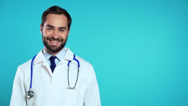Skopiuj kosmiczny portret uśmiechniętego mężczyzny w profesjonalnym medycznym białym płaszczu trzęsącego głową jak gest zgody i zgody. Lekarz odizolowany na tle niebieskiego studia. — Wideo stockowe