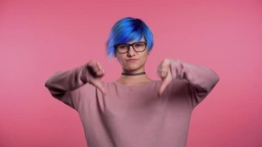 Pembe stüdyo arka planında duran, hoşnutsuzluğunu dile getiren ve kameraya baş parmak hareketi yapan mavi saçlı genç bir kadın. Sevmediğine dair bir işaret taşıyan bir kızın portresi.