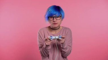 Pretty young girl with blue hairstyle playing video exciting game on Tv with joystick on pink studio wall. Using modern technology.