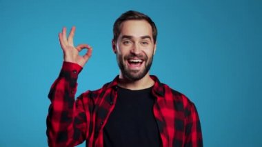 Sakallı genç yakışıklı adam mavi arka planı imzalıyor ve kameraya gülümsüyor..