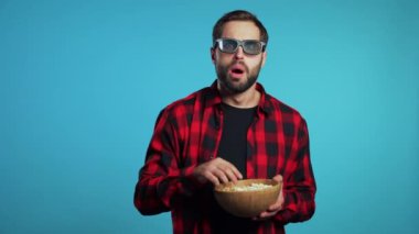 Young man in 3d glasses watching fascinating horror movie and eating popcorn on blue studio background.