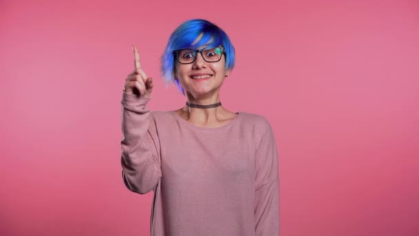 Retrato de una joven mujer pensante con un inusual peinado azul que tiene un momento de idea apuntando con el dedo hacia arriba en el fondo del estudio rosa. Sonriente chica feliz mostrando gesto eureka . — Vídeo de stock