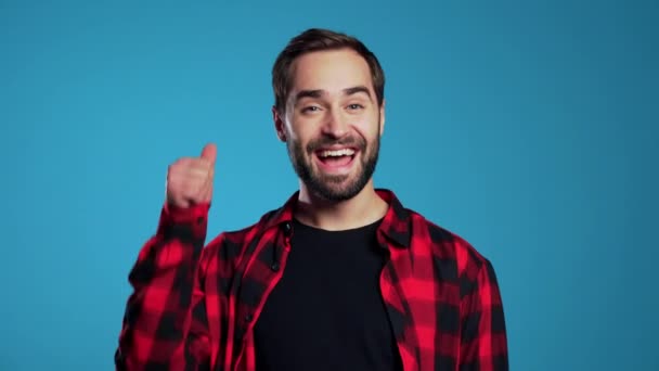Молодий красивий чоловік з бородою робить великі пальці підпис на синьому фоні і посміхається камері . — стокове відео