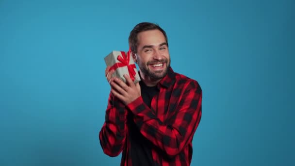 Homem bonito jovem sorrindo e segurando caixa de presente no fundo do estúdio azul. Um tipo com barba . — Vídeo de Stock