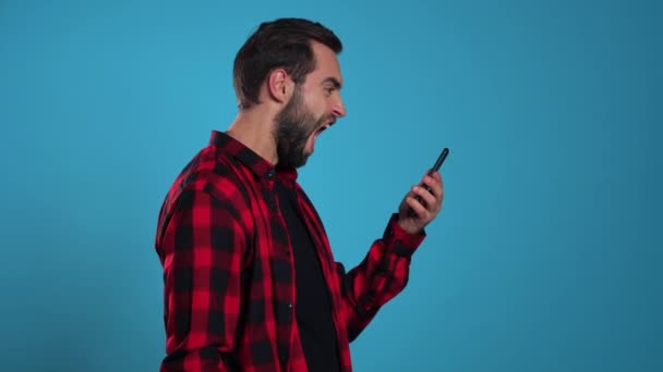 Angry modern hipster in red plaid shirt screaming down his mobile phone. Stressed and depressed man on blue background. — Stock Video