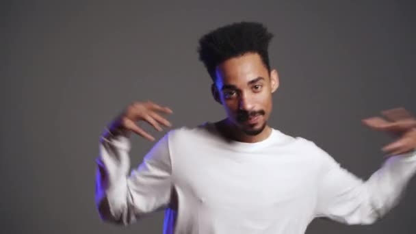 Jonge zeer actieve en energieke Afrikaans-Amerikaanse man in wit dragen glimlachend en dansend in goed humeur op grijze achtergrond. 4k — Stockvideo