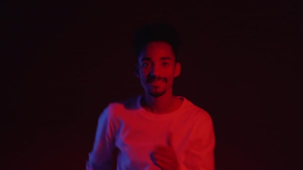 Junger sehr aktiver und energischer afrikanisch-amerikanischer Mann in Weiß, der lächelnd und gut gelaunt auf dunklem Hintergrund mit rotem Neonlicht tanzt. 4k — Stockvideo