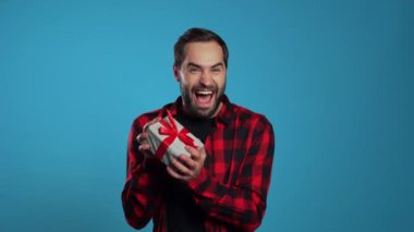 Genç yakışıklı adam gülümsüyor ve elinde mavi stüdyo arka planında hediye kutusu var. Sakallı adam..