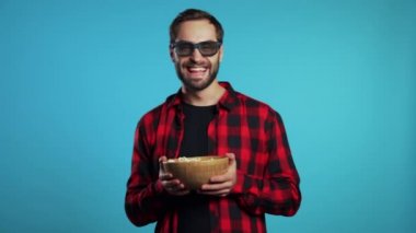 Üç boyutlu gözlüklü genç Avrupalı adam etkileyici bir komedi filmi izliyor, gülüyor ve mavi stüdyo arka planında patlamış mısır yiyor..