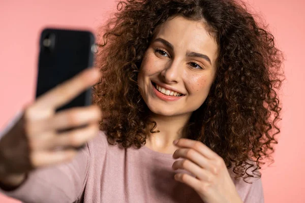 Kıvırcık saçlı, gülümseyen mutlu kız pembe arka planda akıllı telefondan selfie çekiyor. Teknoloji, mobil cihaz, sosyal ağlar kavramı. — Stok fotoğraf