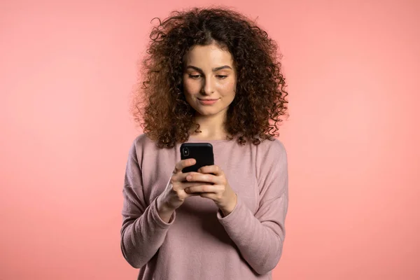 Kıvırcık saçlı, komik makyajlı akıllı telefon kullanan ya da pembe stüdyo geçmişinde internette sörf yapan bir kız. Modern teknoloji - uygulamalar, sosyal ağlar — Stok fotoğraf