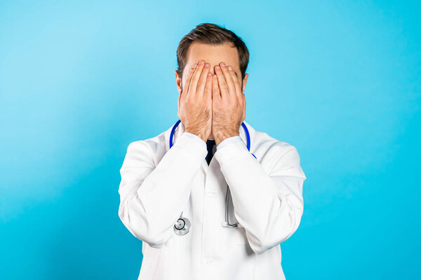 Male doctor covers his face with hand from fatal disappointment, failure. Depressed lost man in professional white coat. Drama, emotions concept.