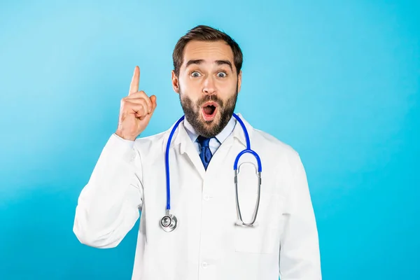 수염을 기르고 있는 의사를 생각하고 있는 젊은 남자의 사진 이 파란 스튜디오 배경을 손가락으로 가리키고 있습니다. 행복 한 의사가 유레카의 몸짓을 하는 모습. — 스톡 사진