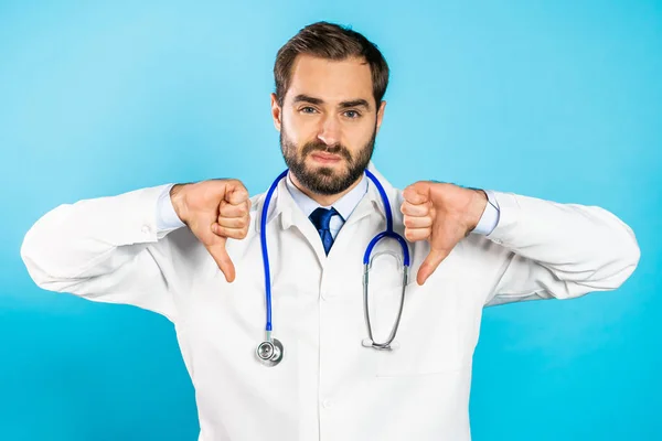 Porträt eines seriösen Arztes im weißen Arztkittel, der eine Geste der Abneigung mit dem Daumen nach unten zeigt. doc man isoliert auf blauem Hintergrund. — Stockfoto