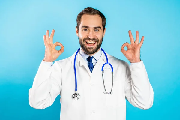 Portret uśmiechniętego lekarza w białym, medycznym płaszczu z oznakowanym gestem Ok. Doc izolowany na niebieskim tle. — Zdjęcie stockowe