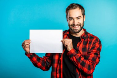 Yakışıklı bir adam elinde yatay beyaz bir a4 kağıt poster tutuyor. Uzayı kopyala Mavi arka planda kırmızı ekose gömlekli gülümseyen hippi..