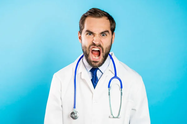 Wściekły lekarz w białym płaszczu krzyczący do kamery. zestresowany i przygnębiony mężczyzna na niebieskim tle. — Zdjęcie stockowe