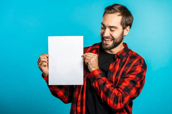 Красивий чоловік тримає вертикальний білий паперовий плакат a4. Копіювати простір. Усміхнений хіпстер у червоній сорочці на синьому фоні . — стокове фото