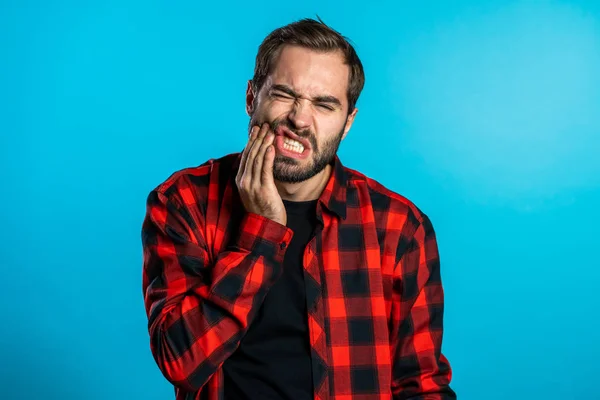 Young handsome man in red shirt with tooth pain on blue studio background. Toothache, dental problems, stomatology and medicine concept. — Stock Photo, Image