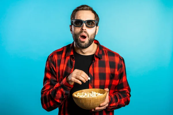 Jeune homme en 3d lunettes regarder film d'horreur fascinant et manger du pop-corn sur fond bleu studio . — Photo