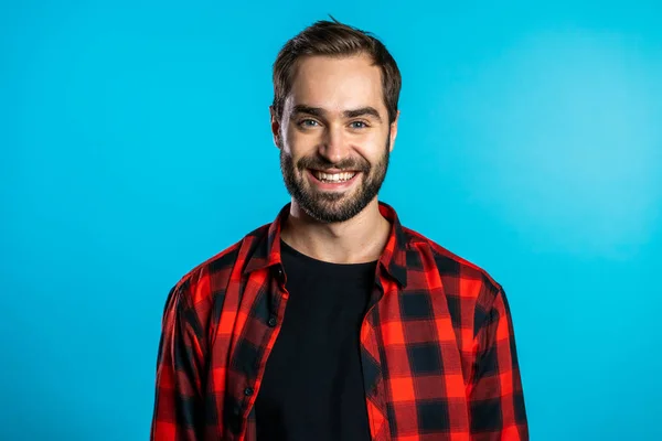 Homem europeu bonito com barba na moda em camisa xadrez vermelho no fundo do estúdio azul. cara alegre sorrindo e olhando para a câmera . — Fotografia de Stock