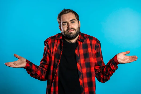 Jovem bonito inseguro cara em vermelho xadrez camisa encolhe os braços, faz gesto de eu não sei, não pode ajudar nada — Fotografia de Stock