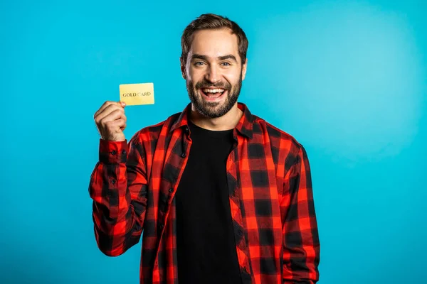 Úspěšný muž v kostkované košili ukazující neomezenou zlatou kreditní kartu na modrém pozadí studia. Student, finanční koncept — Stock fotografie