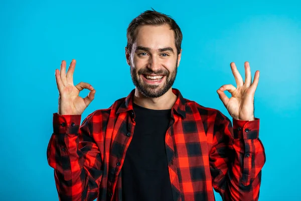 Jeune homme beau avec barbe faisant signe OK sur fond bleu et sourit à la caméra . — Photo