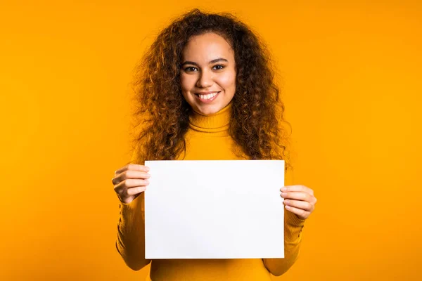 Αρκετά σγουρή γυναίκα κρατώντας οριζόντια λευκή χάρτινη αφίσα a4. Αντιγραφή χώρου. Χαμογελώντας μοντέρνο κορίτσι σε κίτρινο φόντο. — Φωτογραφία Αρχείου