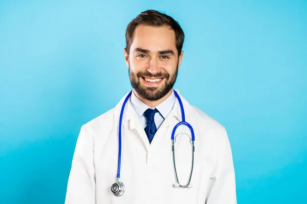 Portret młodego uśmiechniętego mężczyzny w profesjonalnym medycznym białym płaszczu izolowany jest na niebieskim tle pracowni. Lekarz z brodą i stetoskopem — Zdjęcie stockowe