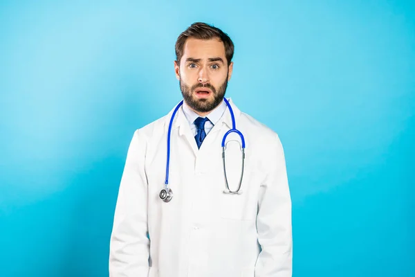 Portret młodego rozczarowanego mężczyzny w profesjonalnym medycznym białym płaszczu odizolowanym na niebieskim tle pracowni. Zapomniany lekarz z brodą i stetoskopem — Zdjęcie stockowe