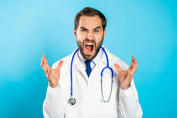 Wściekły lekarz w białym płaszczu krzyczący do kamery. zestresowany i przygnębiony mężczyzna na niebieskim tle. — Zdjęcie stockowe