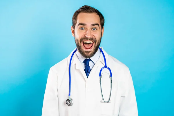 Portret młodego zaskoczonego mężczyzny w profesjonalnym medycznym białym płaszczu izolowany jest na niebieskim tle pracowni. Podekscytowany lekarz z brodą i stetoskopem — Zdjęcie stockowe