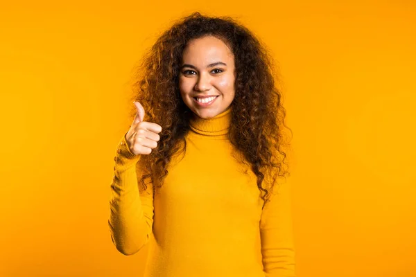 Winner. Success. Positive girl making thumbs up sign over yellow background and smiles to camera. Body language. Young mixed race curly woman